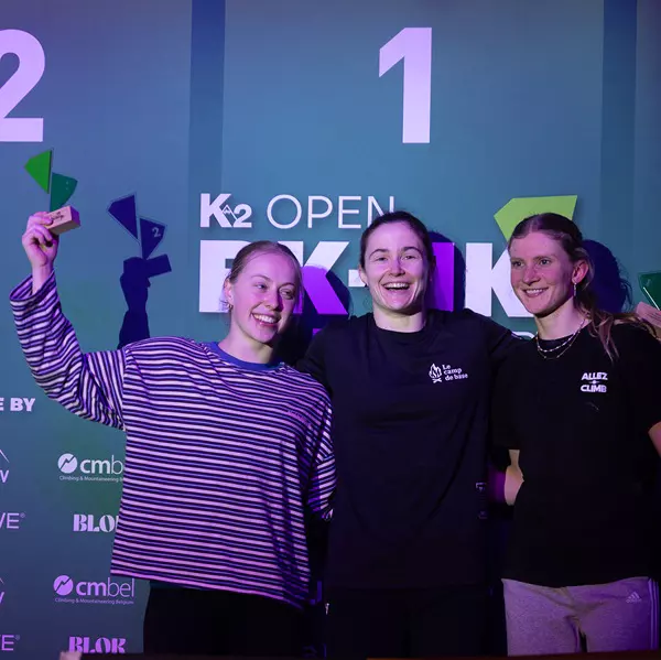 Drie jonge vrouwen staan glimlachend op een podium voor een groene achtergrond waarop ‘K2 Open’ staat geschreven. De vrouw in het midden staat op het hoogste podium, terwijl de anderen groene trofeeën omhoog houden.